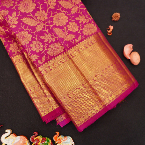 A folded silk saree with a rich magenta pink body and a wide gold zari floral border, displayed on a dark background with decorative elephant and fruit props.