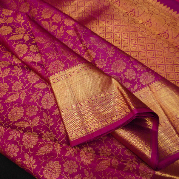 A close-up of a rich magenta pink silk fabric with intricate gold zari floral patterns and a wide gold border.