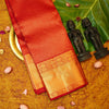 A folded red and gold silk saree is displayed on a textured golden surface, styled with a large green leaf, two small dark figurines, and scattered pink flower petals.