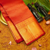 A folded red and gold silk saree with intricate zari embroidery, displayed on a textured golden surface alongside decorative props including small black figurines, a green leaf, and pink flower petals.