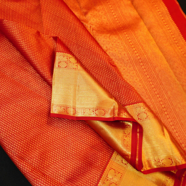 A close-up of an red and gold woven fabric, likely a silk saree or scarf, showing its fine texture, metallic gold zari work, and red border details.