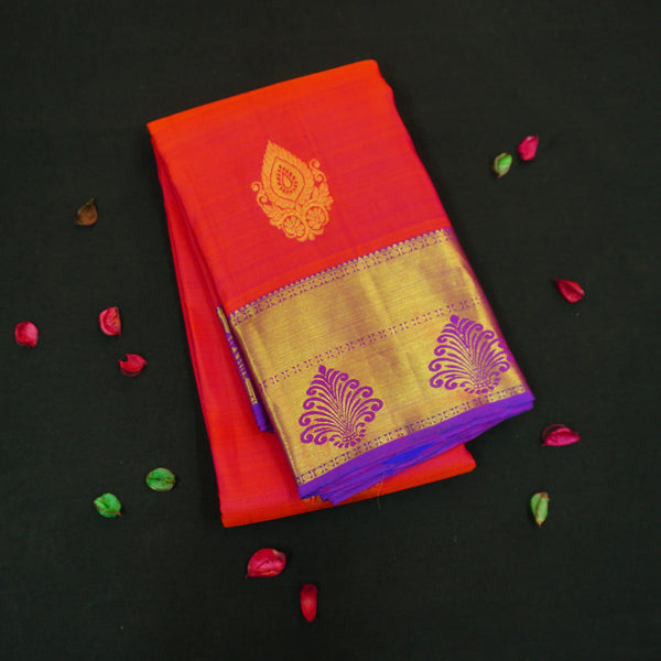Tomato red Kanchipuram silk saree with violet zari border and pallu, featuring gold thilagam motifs.