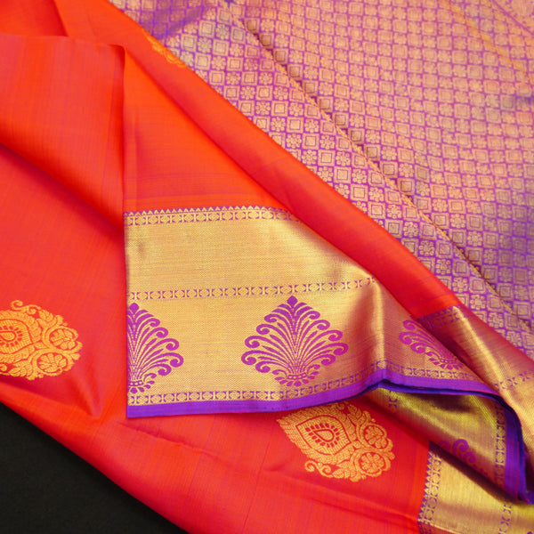 Tomato red Kanchipuram silk saree with violet zari border and pallu, featuring gold thilagam motifs.