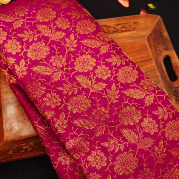 A close-up of a rich magenta pink brocade fabric with an intricate gold floral pattern, draped over a carved wooden tray.