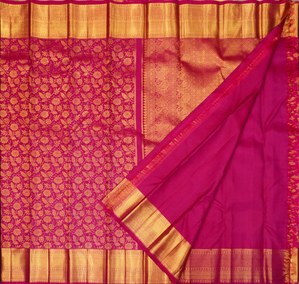 A close-up of a rich magenta pink and gold woven fabric, likely a silk saree, showing a detailed floral pattern on the body, a smooth magenta section, and a wide gold zari border.