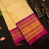 A cream-colored silk saree with a vibrant magenta border and intricate gold zari embroidery, displayed on a black surface with decorative wooden figurines.