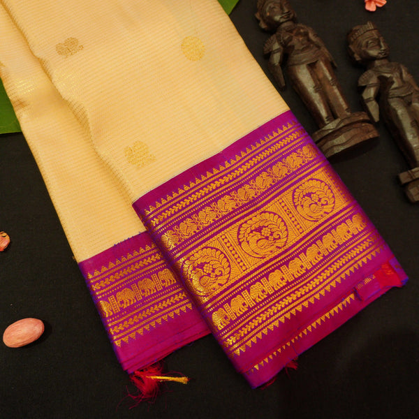 A cream-colored silk saree with a vibrant magenta border and intricate gold zari embroidery, displayed on a black surface with decorative wooden figurines.