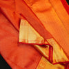 A close-up of an red and gold woven fabric, likely a silk saree or scarf, showing its fine texture, metallic gold zari work, and red border details.